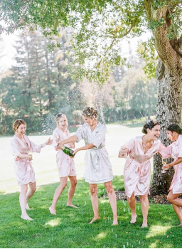 Five women in satin dressing gowns move around excitedly as a bottle of champagne sprays all over outdoors