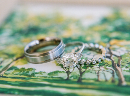 Wedding bands and a rings with white stones sit on a painted surface depicting trees in watercolor