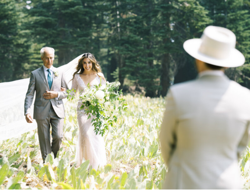 A man walks a bride to a man waiting in a beige suit and sunhat