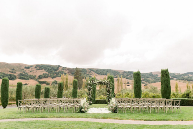 Chairs lined up on the grass overlooking a beautiful hilly terrain and foliage with white petals strewn in the aisle with floral décor and a rectangular arch at the altar