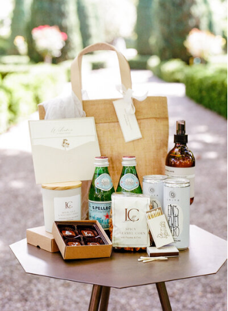 A welcome gift bag arranged with refreshments, chocolates, sparkling water, and locally inspired items, perfect for greeting wedding guests.