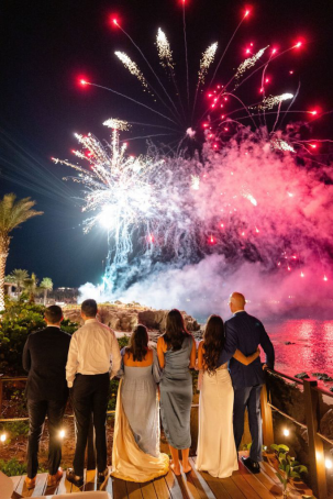 A group of guests watching a breathtaking fireworks display over the ocean at night, with red and white bursts lighting up the sky.