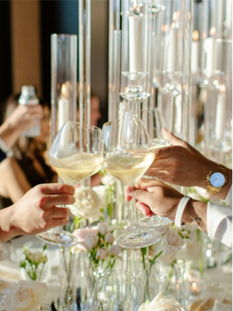 Close-up of hands holding glasses of champagne for a toast, surrounded by tall candles and floral arrangements at a rehearsal dinner.