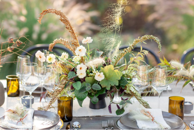 A closeup of a floral decoration with dried stems, fresh flowers, and wild grass, with tableware, runners, and glasses