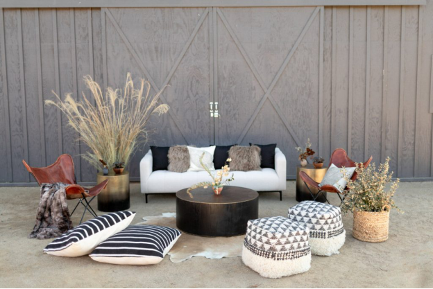 A white sofa with black, grey, and white cushions, black and white striped cushions, bean bag seats, and chairs arranged around a round table on a skin with planter pots with dried grass and stems and a grey door and wall in the back