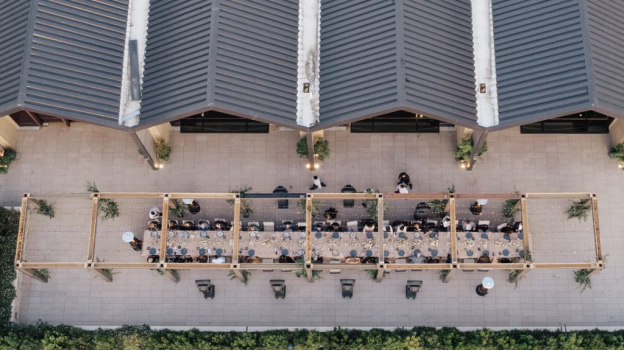 A long table with chairs seated with guests under a canopy of fairy lights outdoors decorated with plates, cutlery, floral decorations, and candles by a building structure