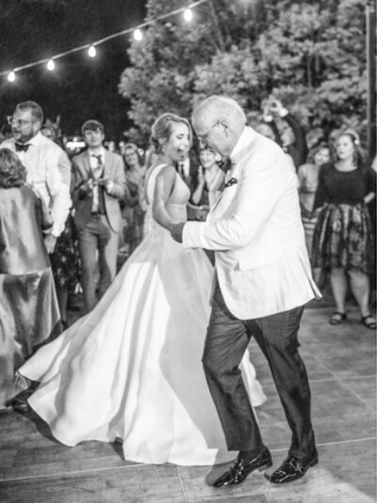 Bride dancing with her father under string lights, surrounded by guests, creating a timeless wedding memory.
