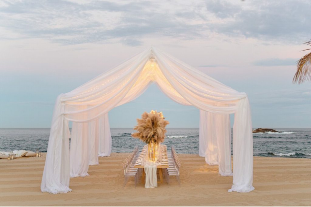 Beachfront luxury dining set up under a white canopy with pampas grass centerpieces.
