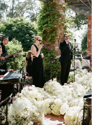  A live band performing at a wedding reception with singers surrounded by lush greenery and white floral arrangements