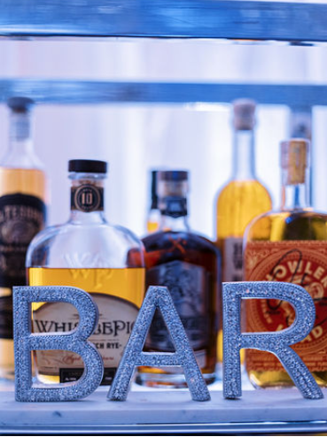 A sparkly silver "BAR" sign sits in front of various liquor bottles on a glass shelf.