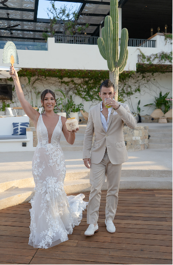 A bride in a lace gown and groom in a tan suit raise drinks in a celebratory toast.