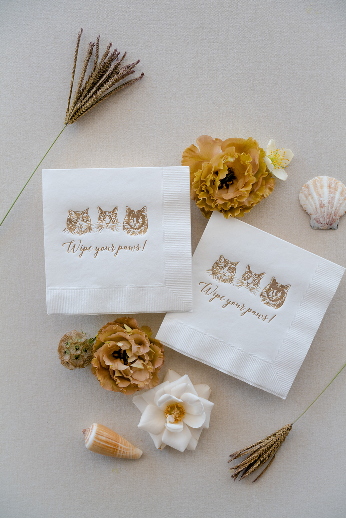 Two white napkins printed with cartoon cats and "Wipe your paws!" are arranged with flowers and seashells on a light surface.