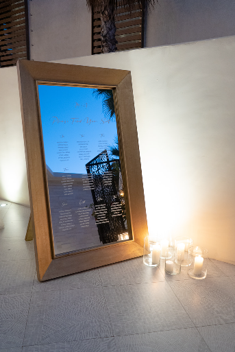 A framed mirror serves as a seating chart with guest names and table numbers, illuminated by nearby candles.