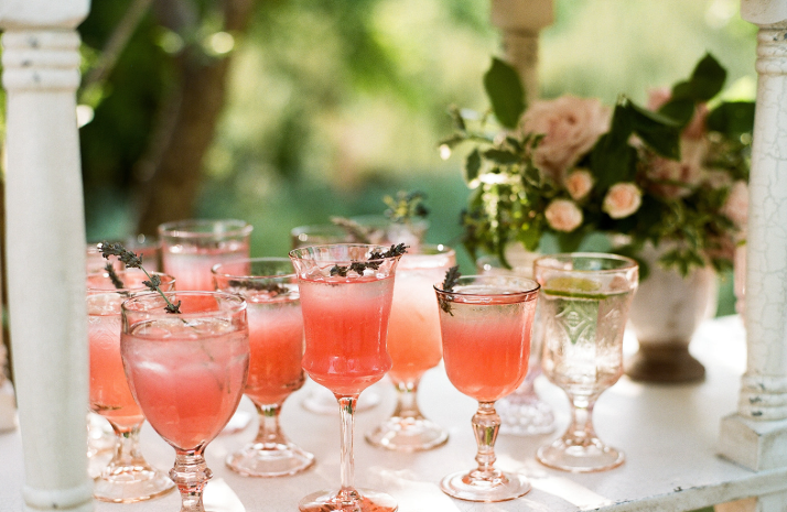 Several glasses filled with pink grapefruit cocktails, garnished with herbs, are arranged on a white surface with flowers.