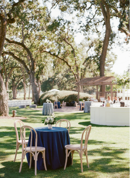 Outdoor wedding cocktail lounge with navy table linen and wooden chairs under oak trees