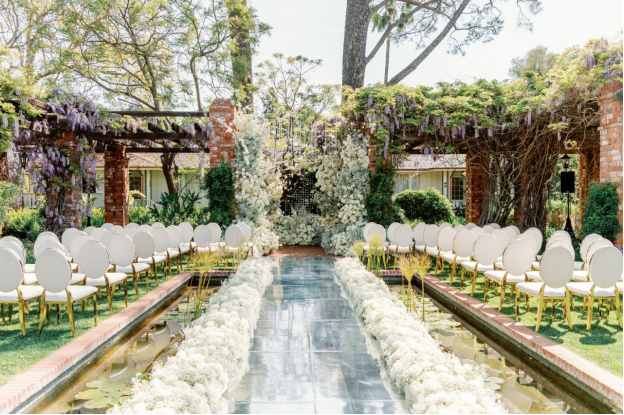 Romantic garden wedding ceremony setup with white floral aisle and wisteria-covered pergola