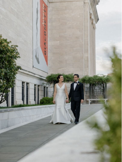 Bride and groom walking hand in hand outside a historic New York building with tall stone walls and museum banners.