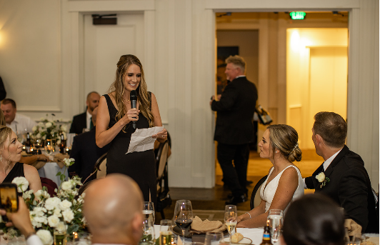 Maid of honor delivering a wedding speech indoors while the bride and groom listen.
