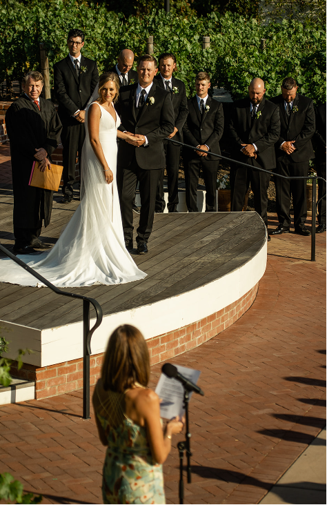 Bride and groom holding hands during wedding vows on an outdoor stage with attendants.